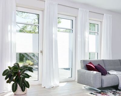 Lichtdurchlässige Plissee in Weiß angebracht in bodentiefen Fenstern in einem Wohnzimmer Lichtdurchlässige Plissee in Weiß angebracht in bodentiefen Fenstern in einem Wohnzimmer
